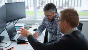 Two business advisors, Simon Ross and Aaron Cassidy, reviewing financial documents together at a desk in a modern office, discussing strategy while working on a computer and handwritten notes.
