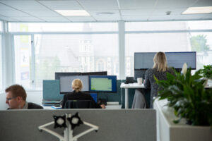 Modern office workspace with employees working at desks and standing desks, using multiple monitors in a bright, open-plan environment with large windows and indoor plants.