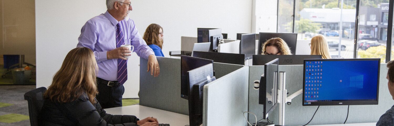 Team working in a bright, open-plan office with multiple desks and monitors, while a senior colleague stands nearby holding a coffee and speaking with staff.